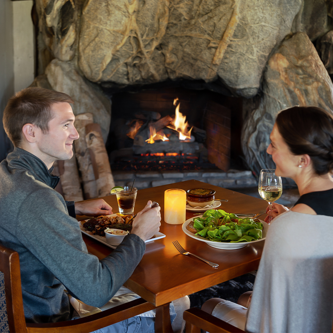 A couple talking over dinner beside a blazing fire in the glacial-boulder fireplace, with ribs, a soup crock, salad and a wine glass etched "Stafford's"