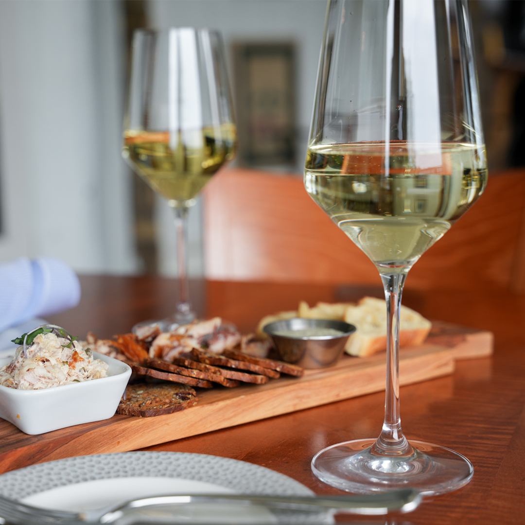 Two glasses of white wine on a sunlit wood table beside a cherry board of whitefish dip, smoked fish, rye crisps, baguette and a dipping cup