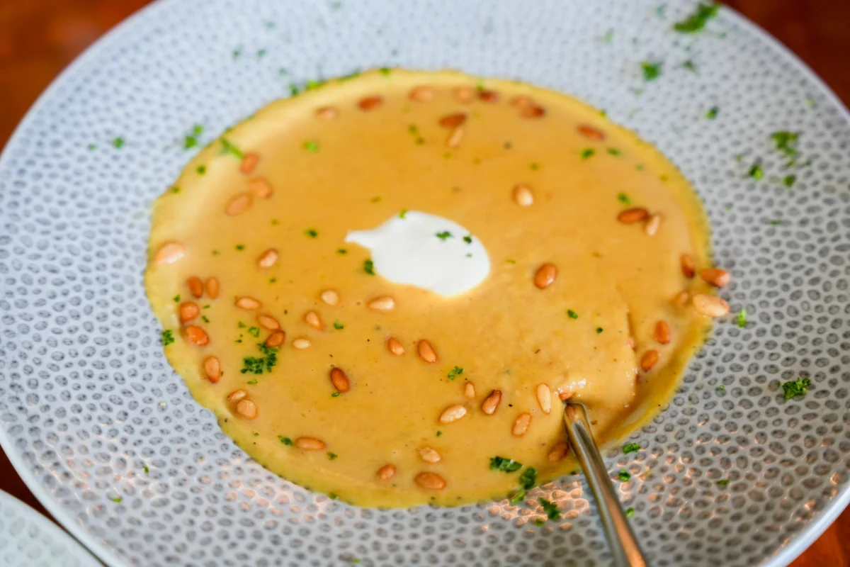 Creamy golden soup topped with toasted pine nuts, chopped parsley, and a dollop of crème fraîche in a speckled bowl with a spoon