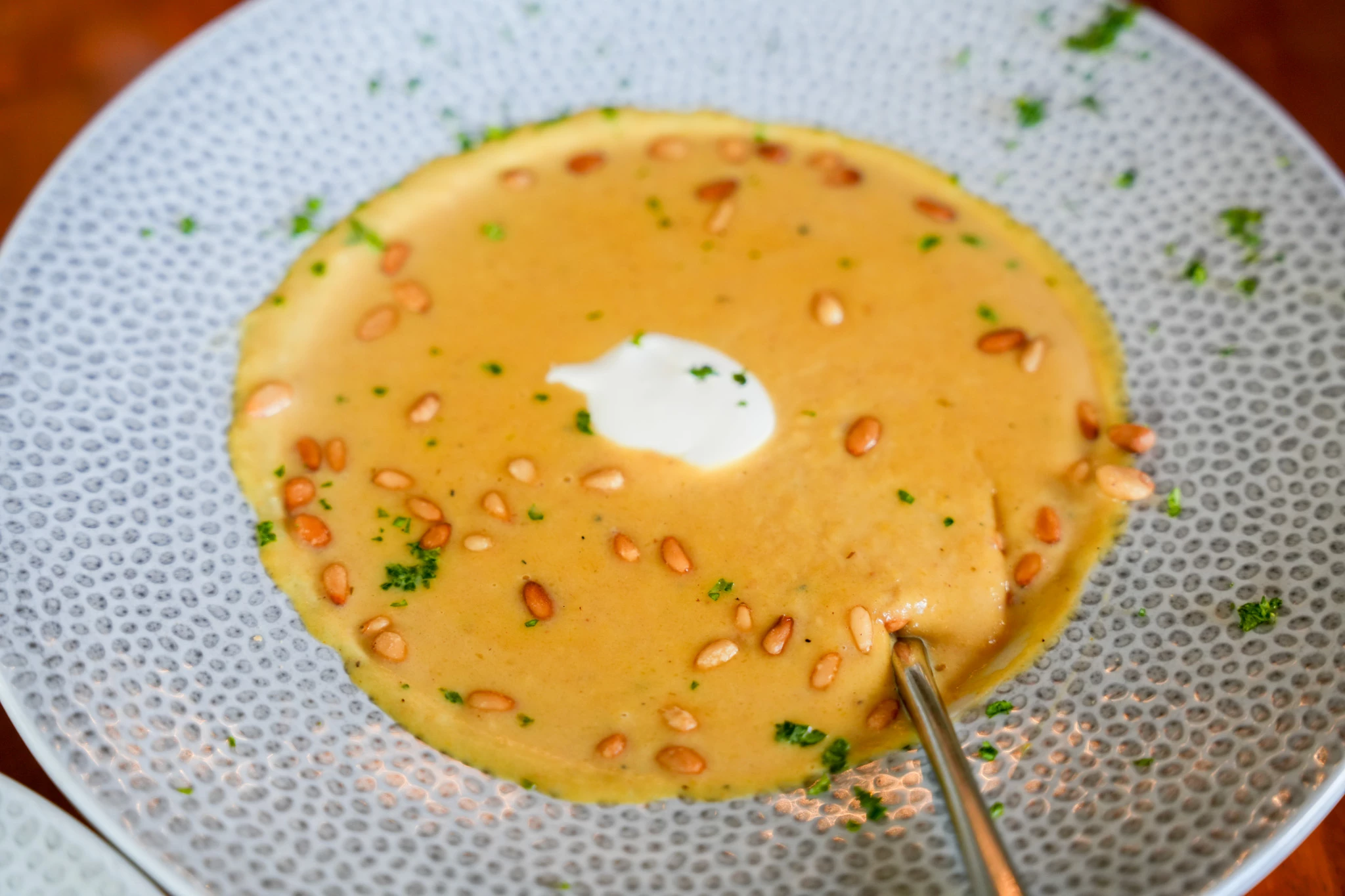Creamy golden soup topped with toasted pine nuts, chopped parsley, and a dollop of crème fraîche in a speckled bowl with a spoon