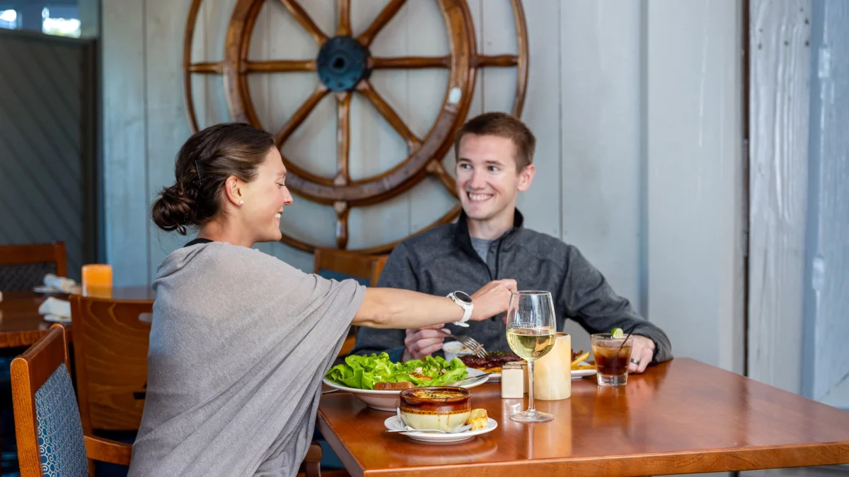 A woman and a man smiling across a wood table with a butter-lettuce salad, French onion soup, white wine, and an iced cocktail, beneath a large wooden ship's wheel on the wall