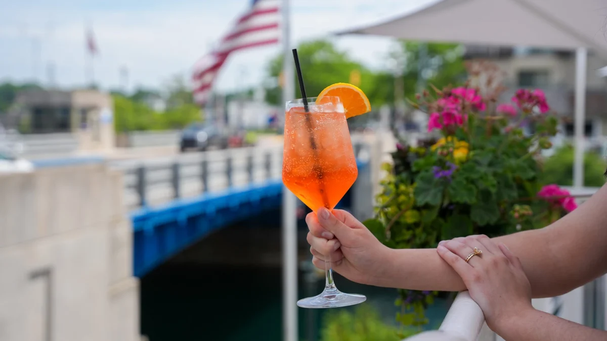 A hand holding an orange spritz garnished with an orange slice above the channel, with the blue drawbridge, an American flag, and pink geraniums behind