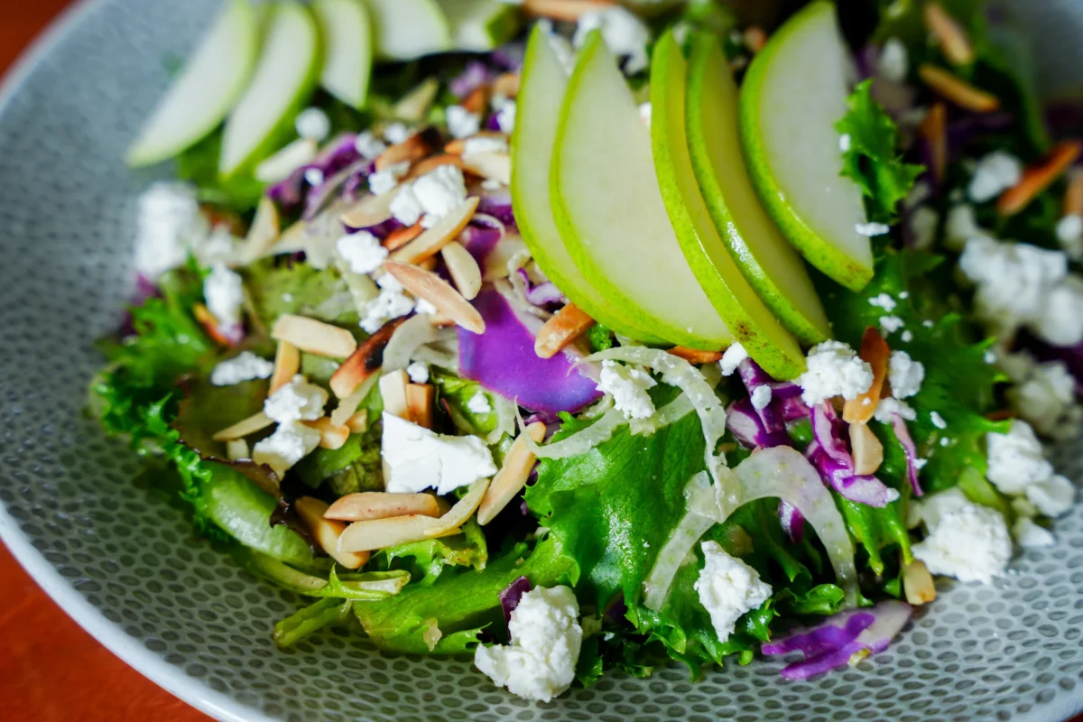 Mixed-greens salad with fanned green apple slices, crumbled goat cheese, toasted slivered almonds, shaved fennel and red cabbage in a grey bowl