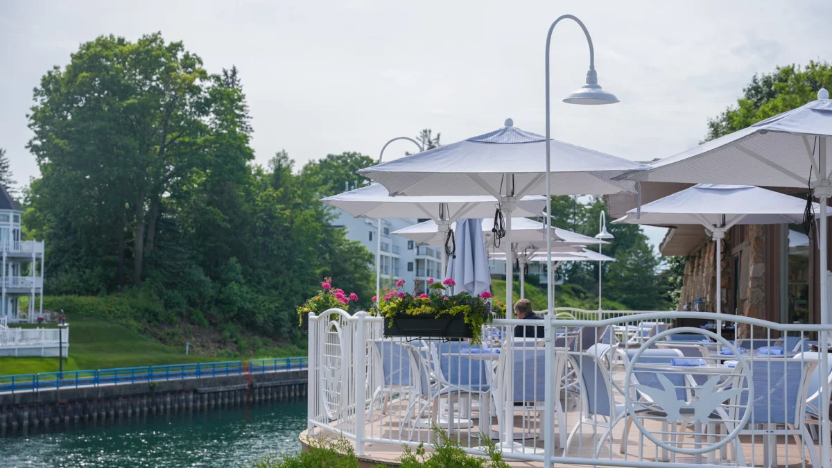 Blue-and-white striped umbrellas over set tables on the waterfront patio, with flower boxes of pink geraniums along the white railing at the channel's edge