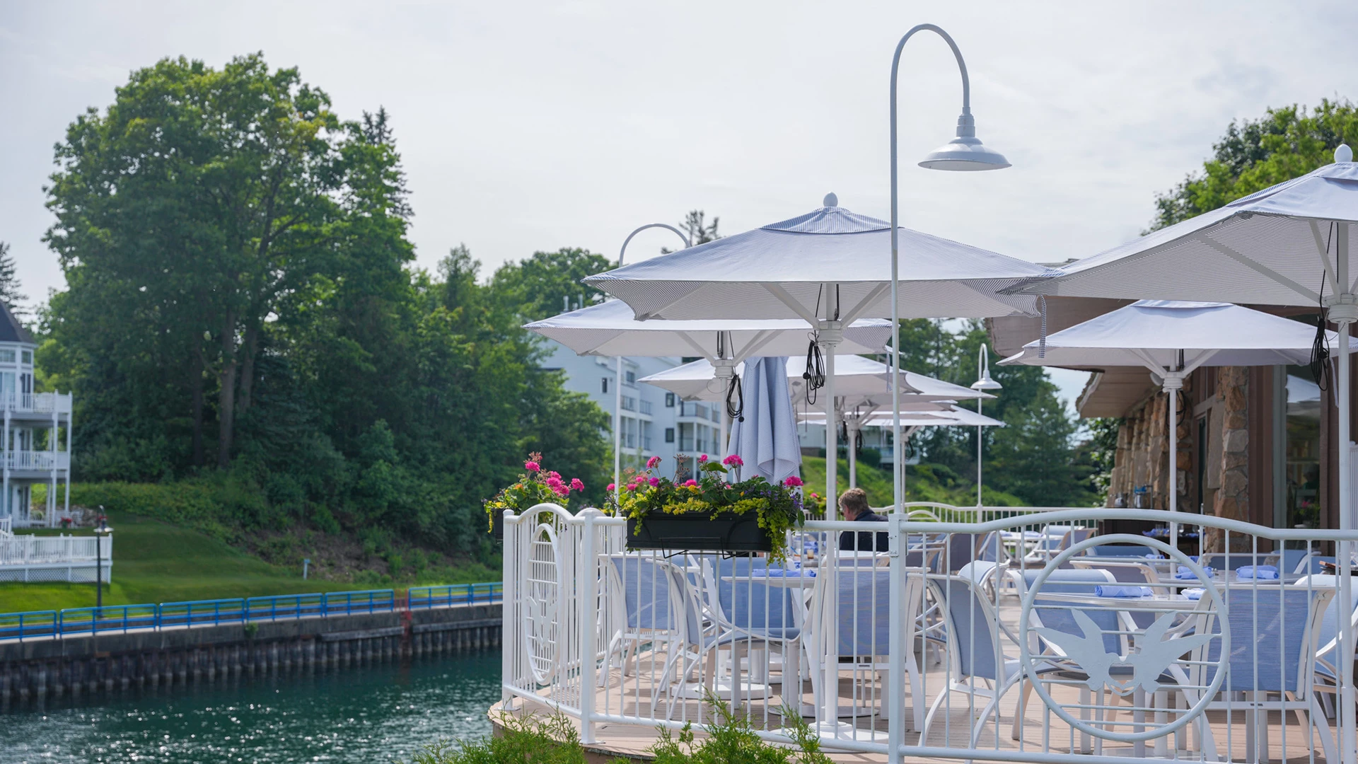 Blue-and-white striped umbrellas over set tables on the waterfront patio, with flower boxes of pink geraniums along the white railing at the channel's edge