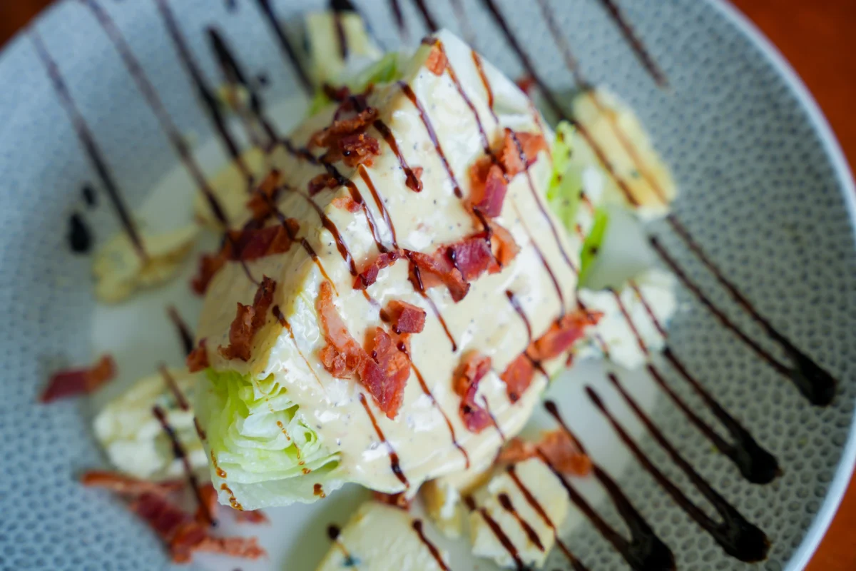 Iceberg wedge salad topped with blue cheese dressing, crumbled bacon, and a balsamic drizzle on a speckled plate