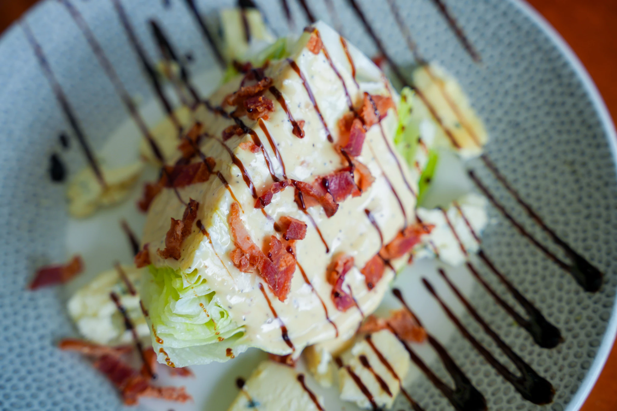 Iceberg wedge salad topped with blue cheese dressing, crumbled bacon, and a balsamic drizzle on a speckled plate
