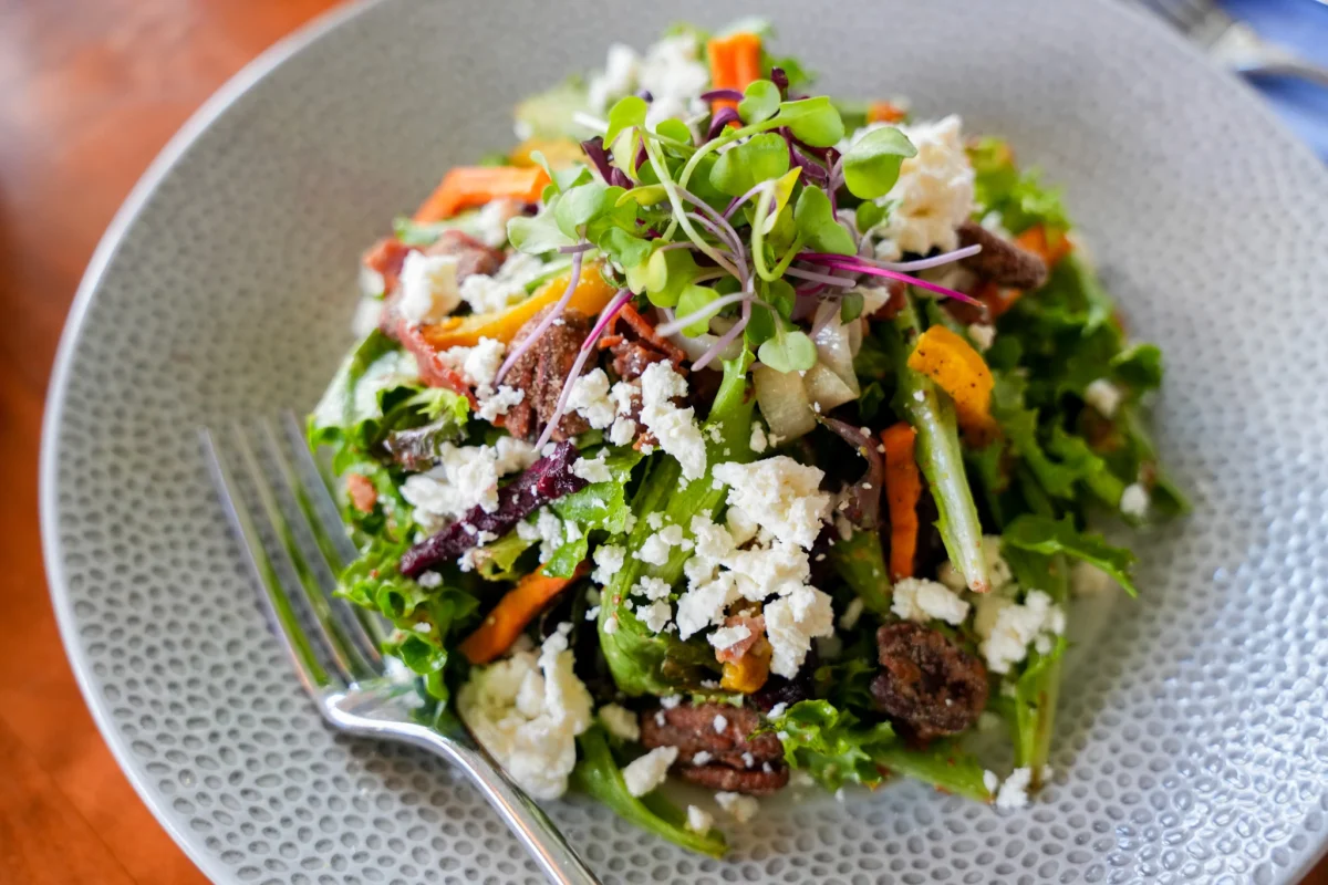 Mixed greens salad with feta crumbles, candied pecans, shaved carrots, beets, asparagus, and microgreens on a speckled bowl with a fork