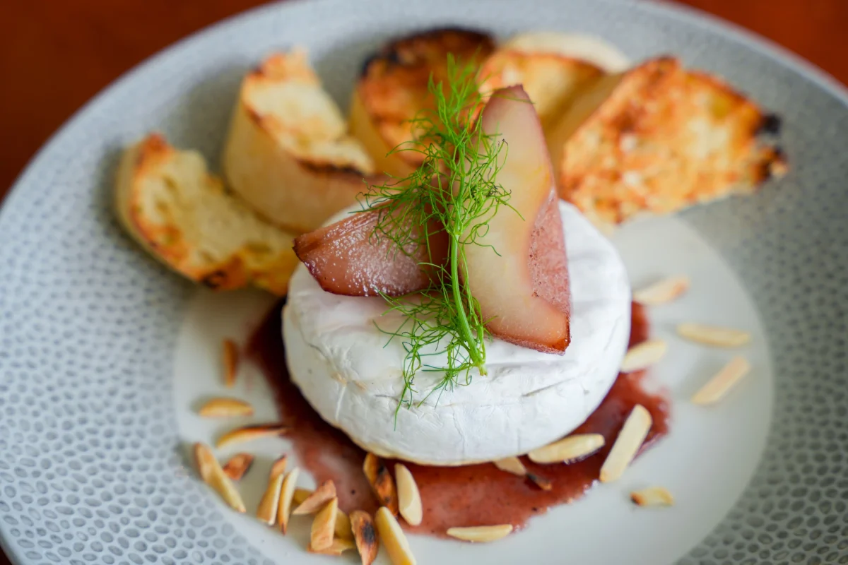 Baked brie wheel topped with wine-poached pear slices and fennel fronds, ringed by red wine sauce, slivered almonds and grilled baguette toasts