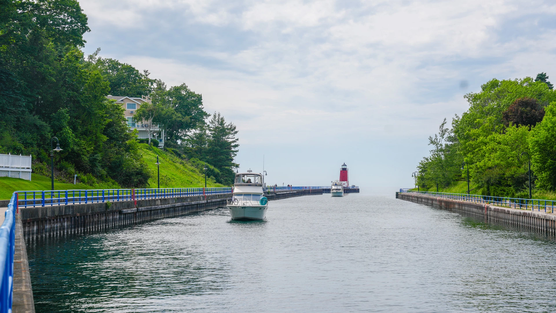 Motor yachts cruising down the Pine River Channel toward Lake Michigan, past the red South Pier lighthouse and blue-railed piers lined with trees