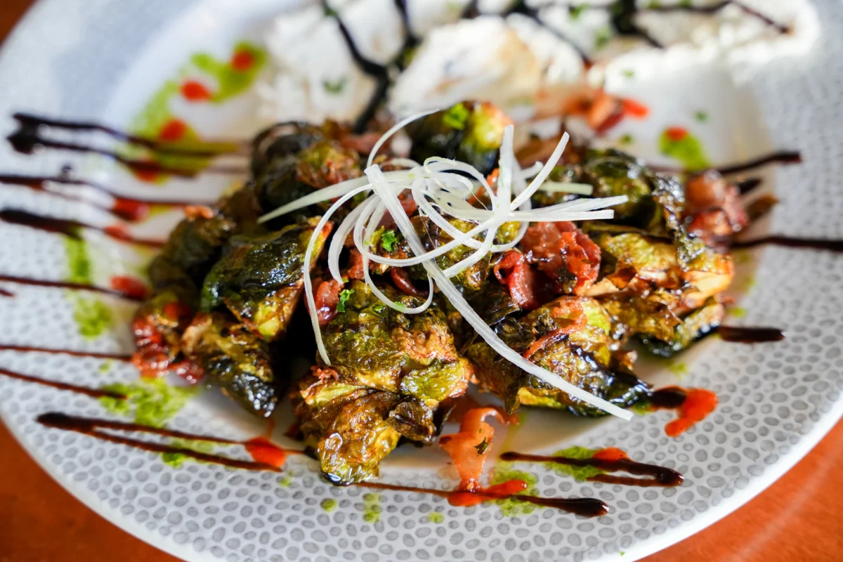 Crispy fried Brussels sprouts with bacon and shaved onion curls, on a dotted white plate drizzled with balsamic, chili, and herb oil