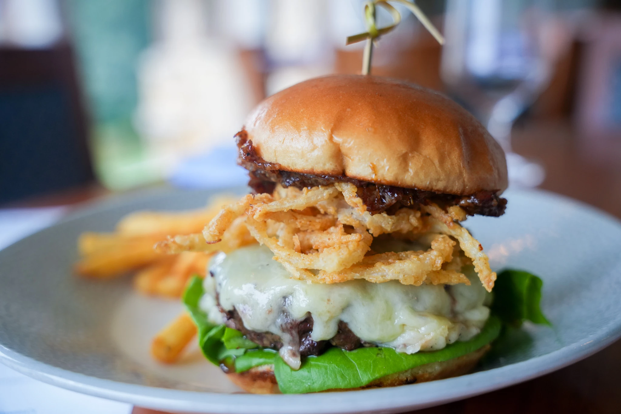 Burger on a brioche bun with melted white cheese, crispy fried onion straws and lettuce, skewered with bamboo and served with fries