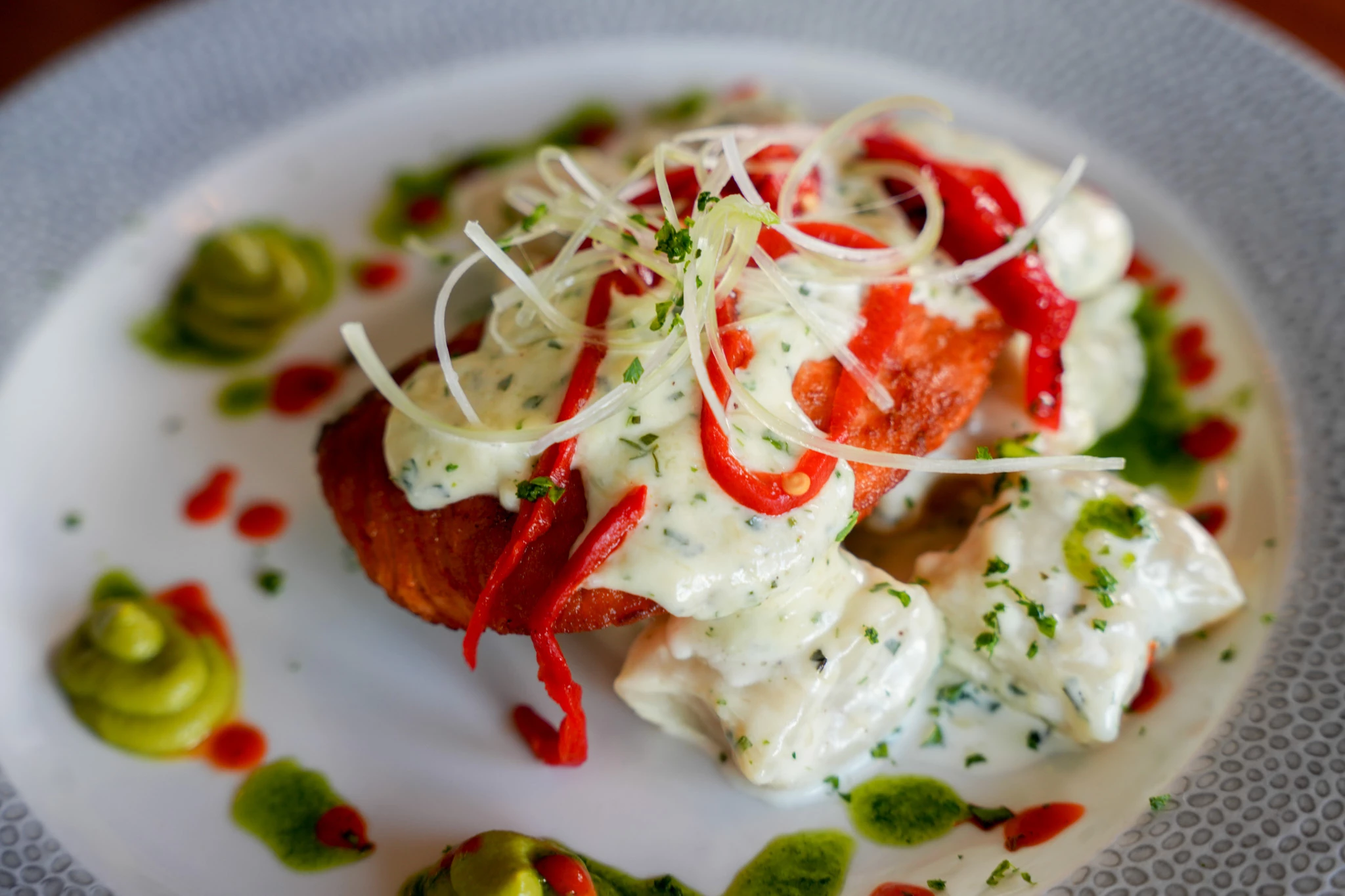 Crisp-fried fish topped with creamy herb sauce, roasted red pepper strips, and shaved onion curls, plated with avocado dollops and green herb oil