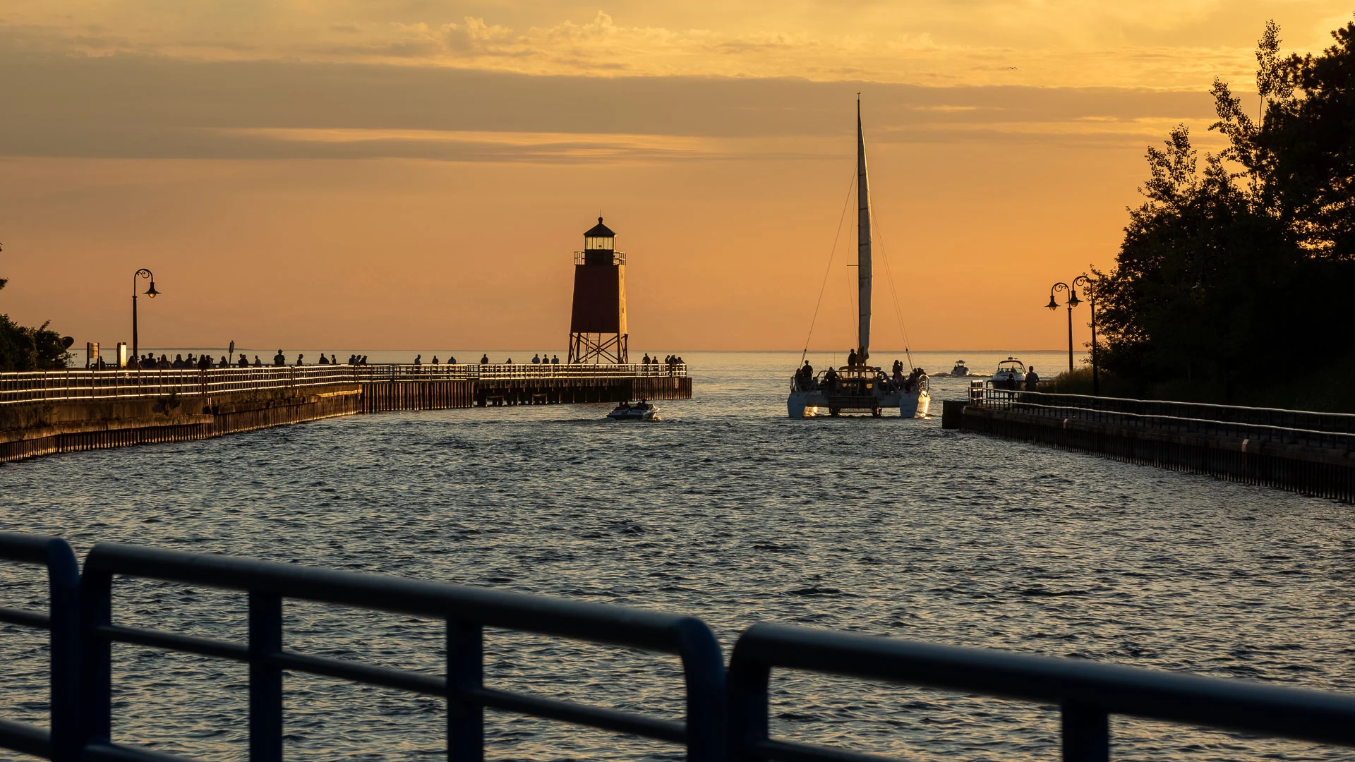 A sailboat and small powerboats heading out the Pine River Channel at sunset, past the red Charlevoix South Pier lighthouse and people gathered along the pier