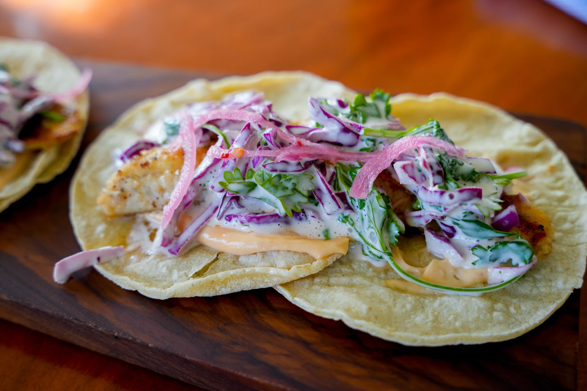 Two fish tacos on corn tortillas with cabbage slaw, pickled red onion, cilantro, and chipotle crema, on a dark wooden board