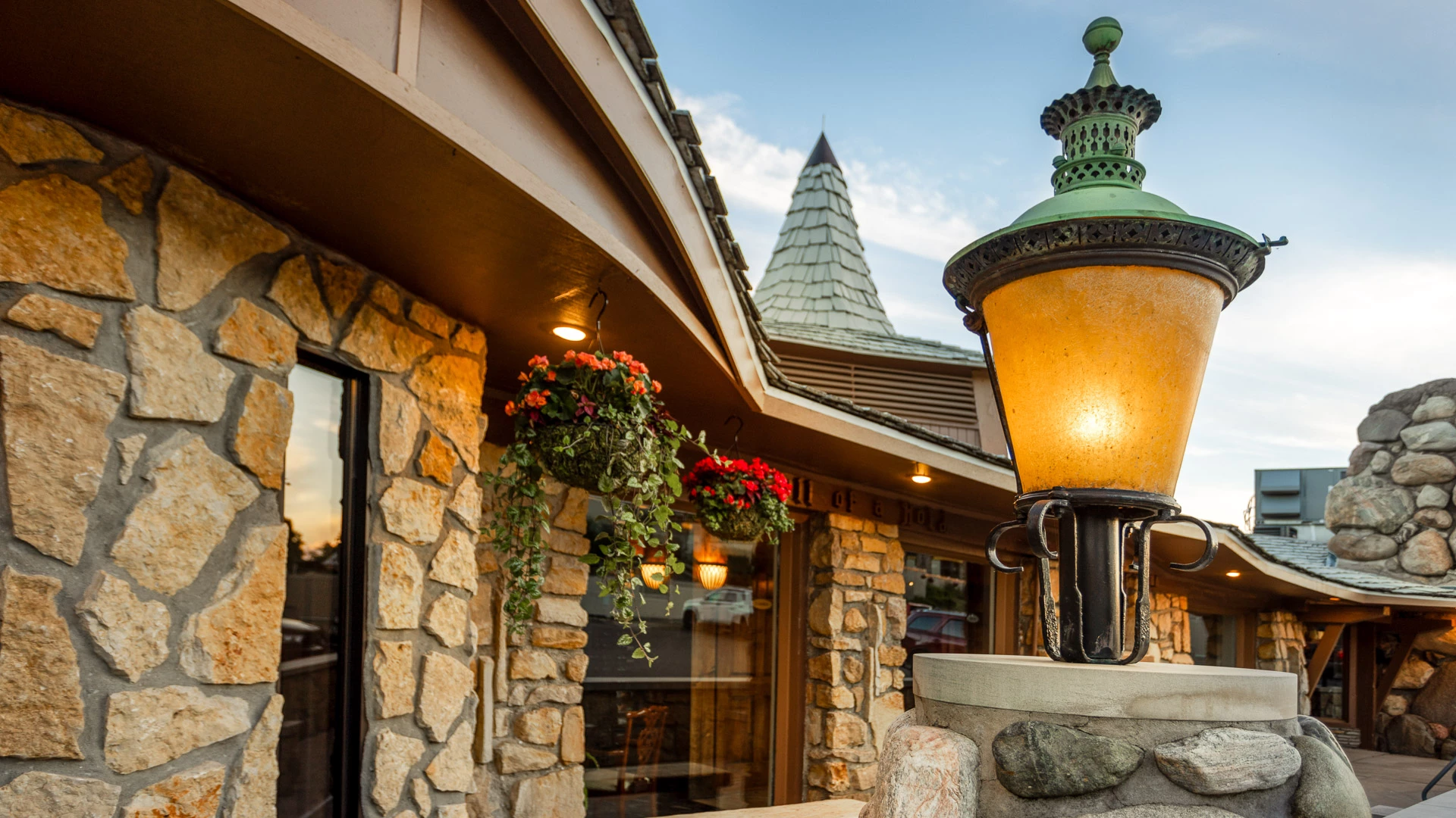 Amber glass lantern on a fieldstone pillar outside the Weathervane at dusk, beside a fieldstone wall, hanging geraniums and a beam carved "Hell of a Hole"