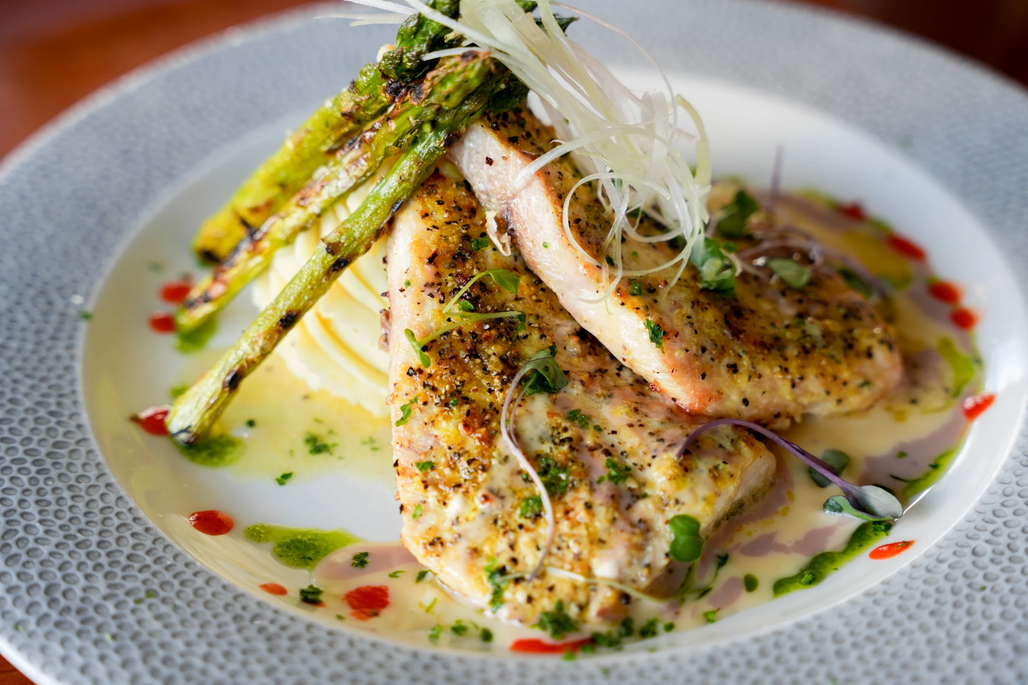 Two pepper-crusted whitefish fillets over mashed potatoes with grilled asparagus, butter sauce, crispy leeks, microgreens and dots of red pepper