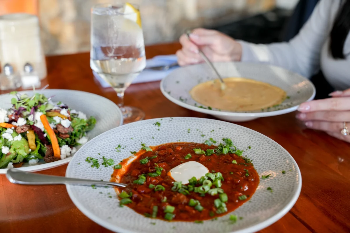 Bowl of red chili topped with sour cream and scallions on a wooden table, with a mixed greens salad with goat cheese, a lemon water glass, and a diner spooning bisque