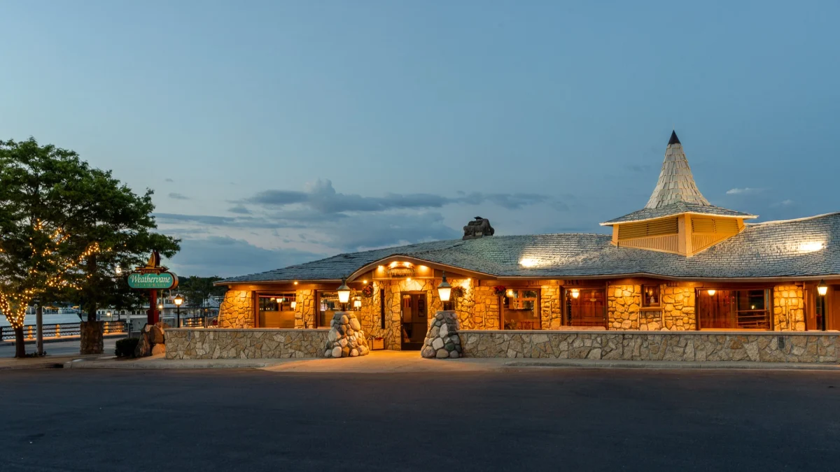 The Weathervane's fieldstone building lit at dusk under its conical shingled turret, with the sailboat road sign "Stafford's Weathervane Restaurant", the entrance sign "Stafford's The Weathervane Restaurant", and "Hell of a Hole" carved on a beam