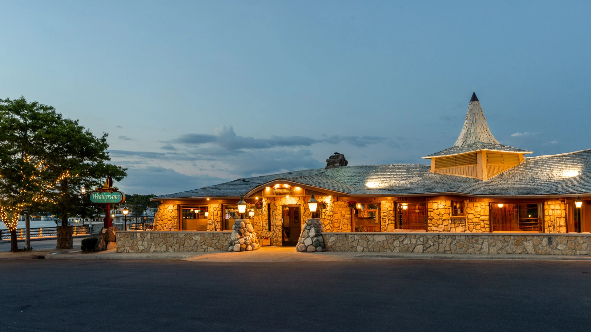 The Weathervane's fieldstone building lit at dusk under its conical shingled turret, with the sailboat road sign "Stafford's Weathervane Restaurant", the entrance sign "Stafford's The Weathervane Restaurant", and "Hell of a Hole" carved on a beam