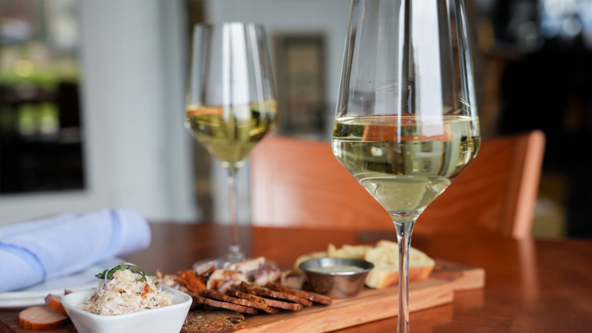 Two tall glasses of white wine beside a cherry serving board of whitefish dip, smoked fish, rye crisps, sliced baguette and a dipping cup, napkin folded alongside