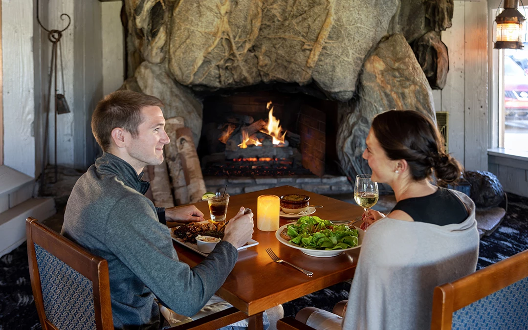 A couple at a wooden table smiling at each other in front of a lit fire under the massive rough-hewn stone hearth, with a candle, a green salad, and a glass of white wine between them