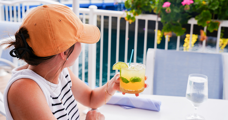 A woman in an orange ball cap and striped tank top holds a citrus cocktail with lime and mint at a white deck table, looking out over the water past a white railing and pink flowers