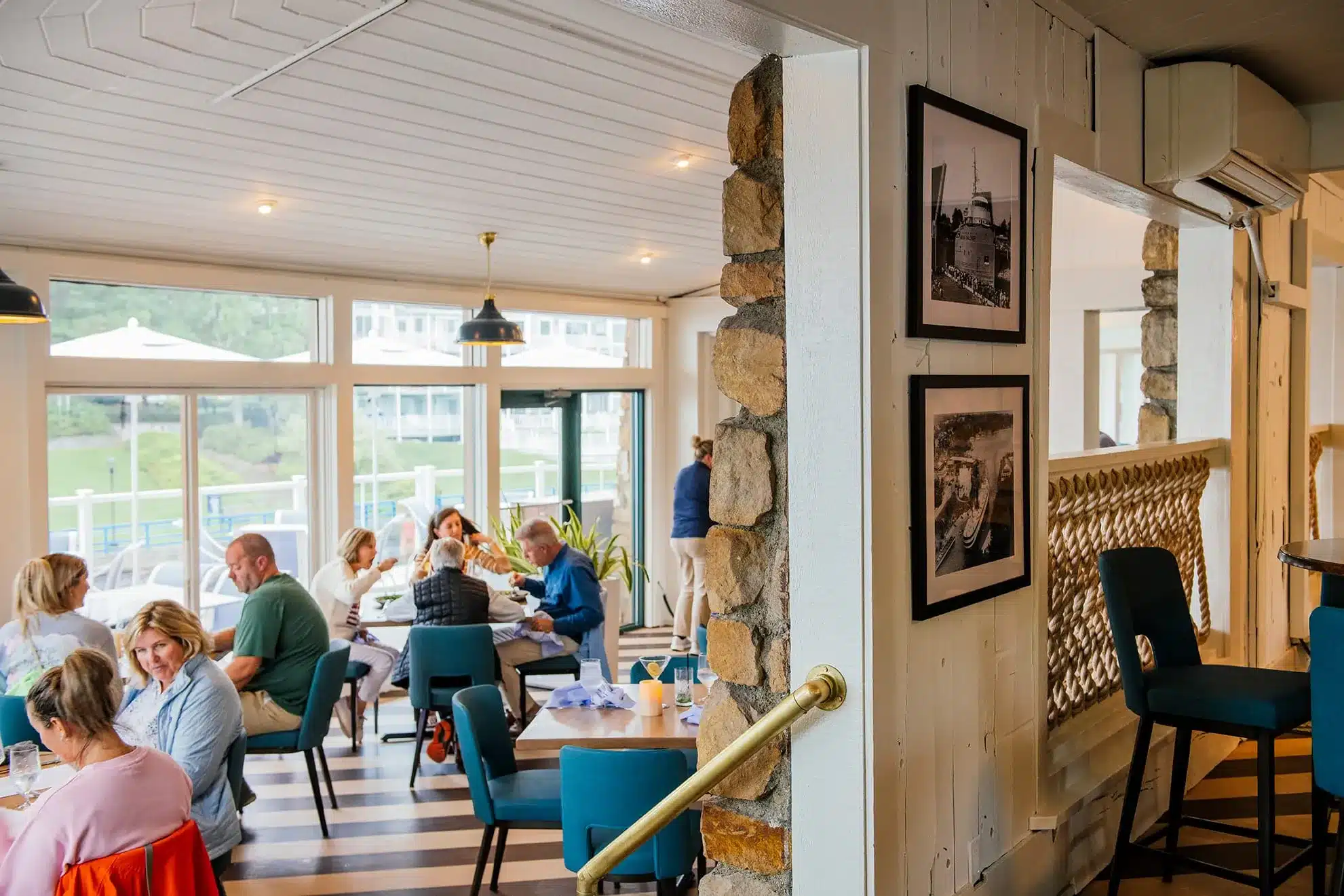 Guests dining beside a fieldstone pillar in the Weathervane's white-planked dining room, with teal chairs, black-and-white nautical photos, rope railing and windows overlooking the channel