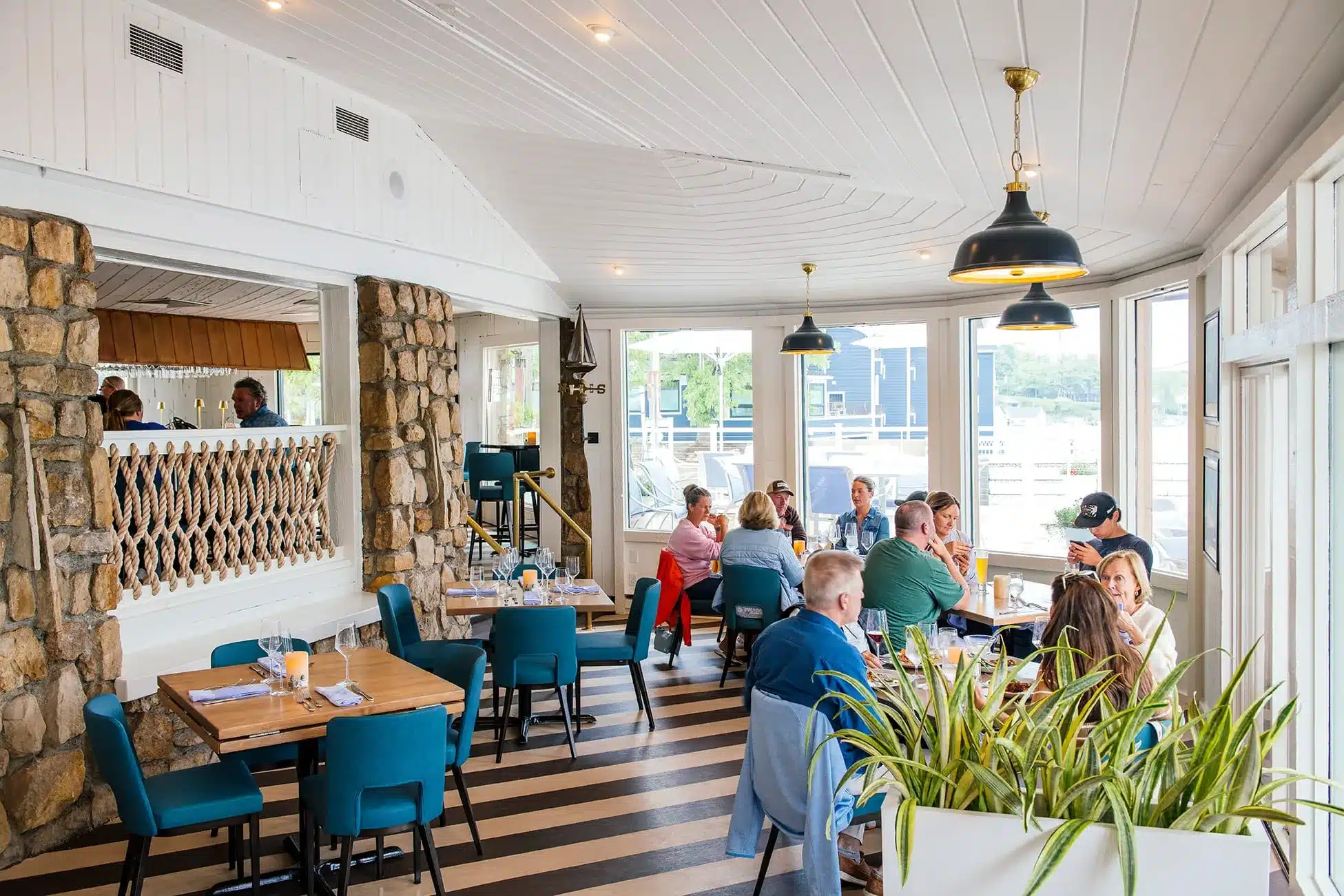 Guests at tables with teal chairs on a black-and-white striped floor in the Weathervane's dining room, under brass pendant lights, fieldstone pillars and a rope railing, with windows onto the waterfront