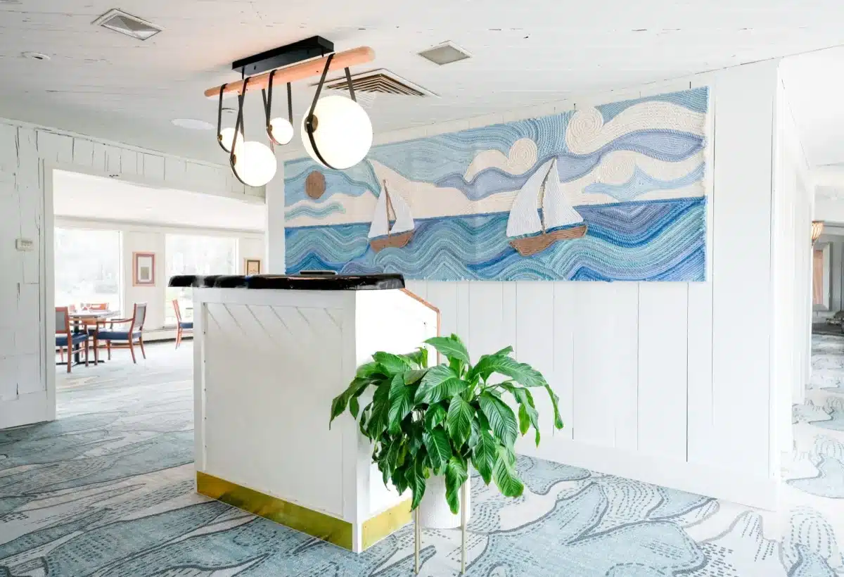White host stand with a potted plant beneath a textured sailboat-and-waves wall hanging and globe pendant lights, with the dining room beyond