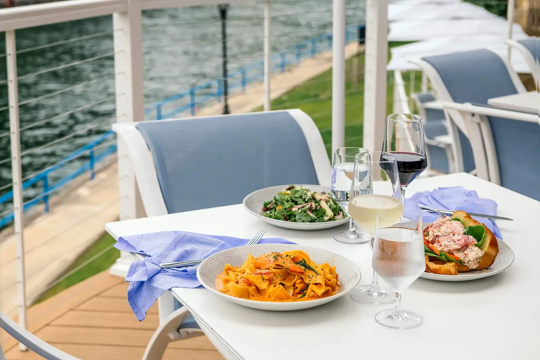 Pappardelle in tomato cream sauce, a lobster roll and an arugula salad with glasses of white and red wine on a patio table overlooking the Pine River Channel