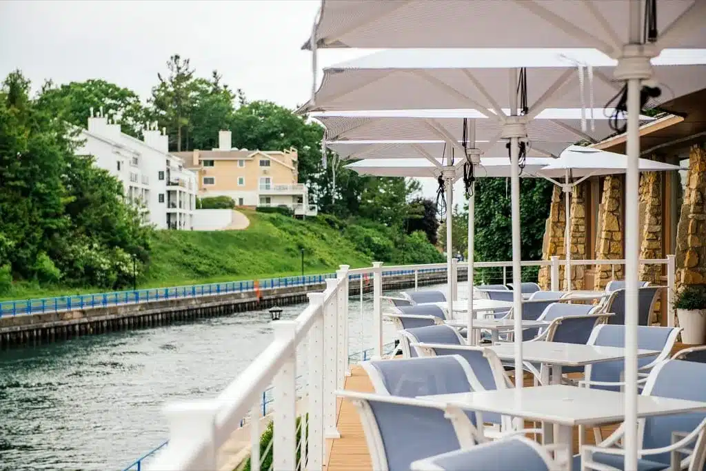 Blue-and-white patio chairs under white market umbrellas along the railing of the Weathervane's waterfront deck, beside the fieldstone building and the Pine River Channel