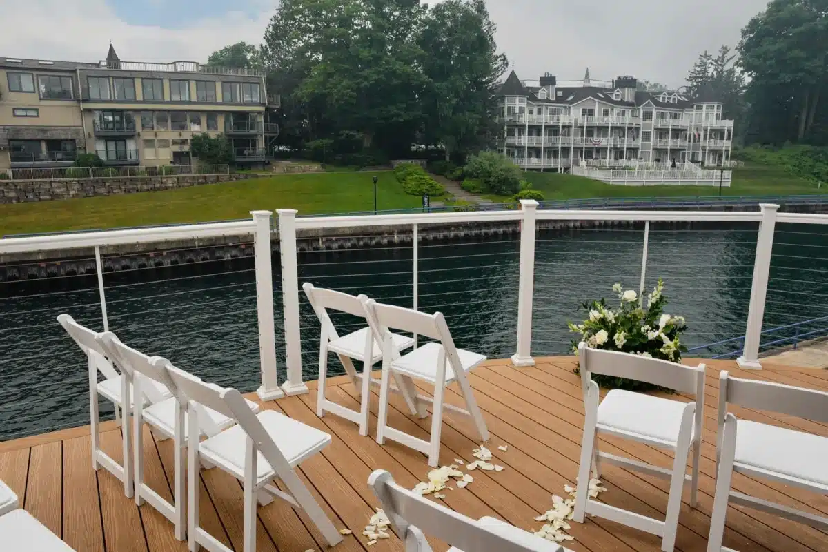 White folding chairs and a bouquet of white roses set up for a wedding ceremony on the Weathervane's waterfront deck, with rose petals on the boards and the Pine River Channel beyond