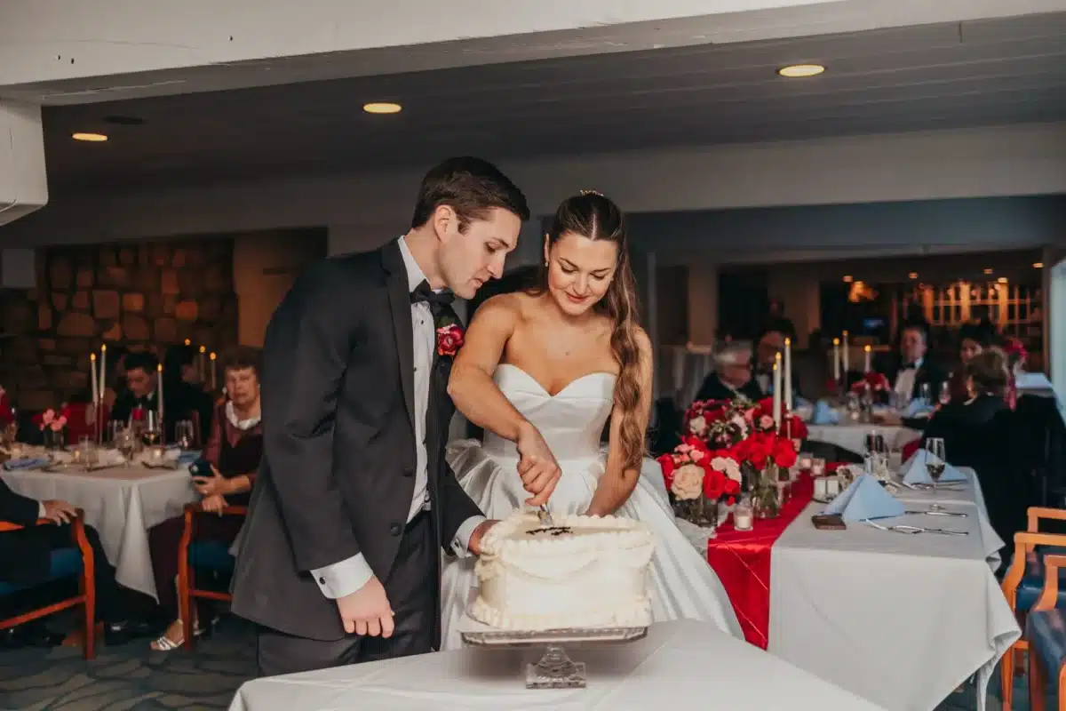 Bride and groom cutting a white wedding cake at the Weathervane, with guests at candlelit tables decorated with red roses behind them